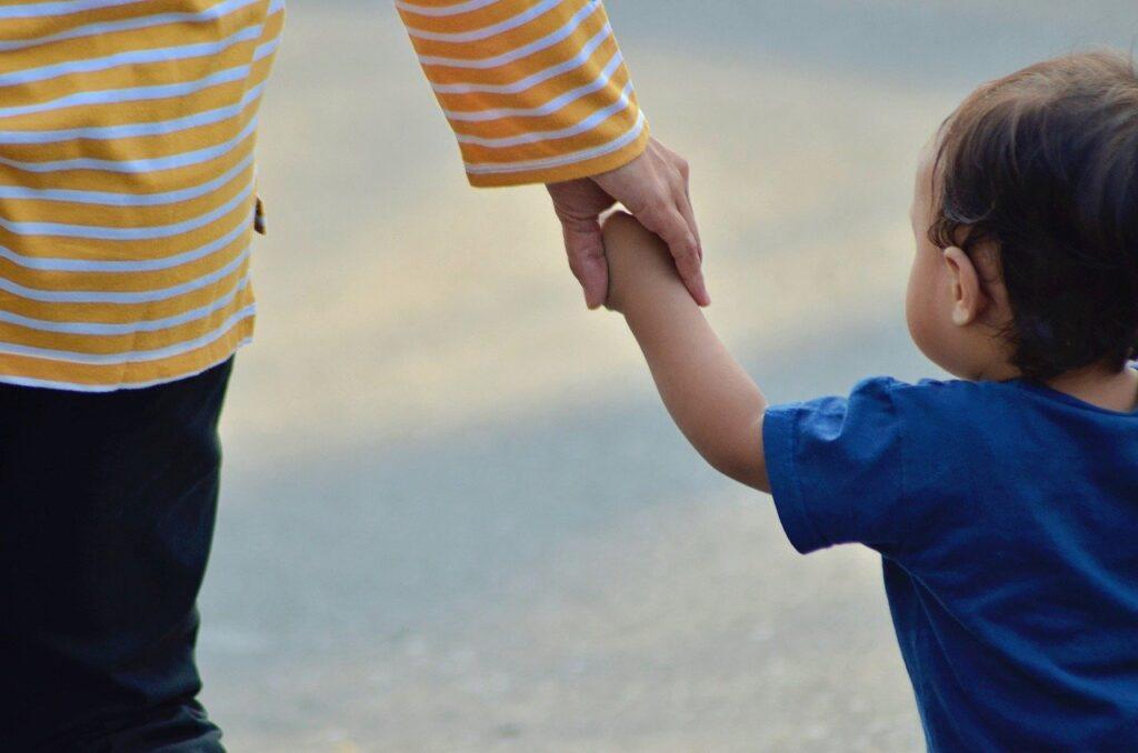 A young child holds their parent's hand as they both walk down the beach.