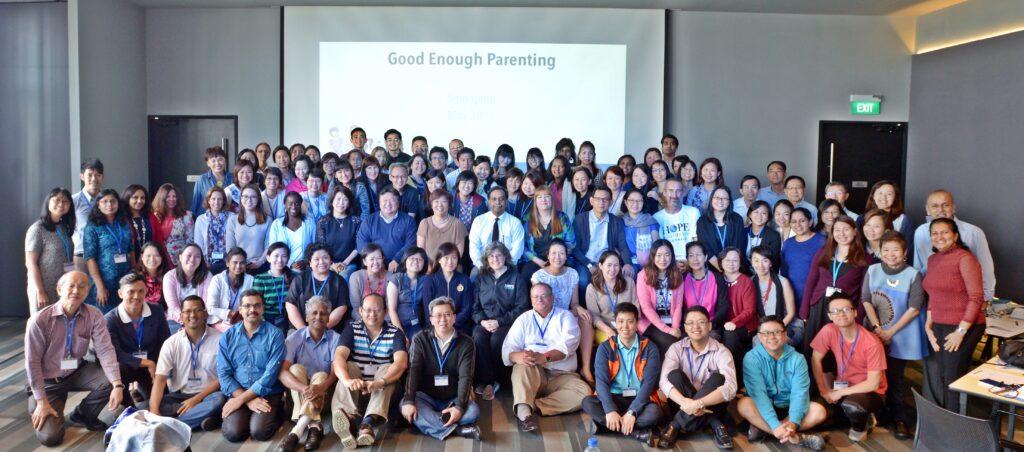 Group photo of about 100 people from various backgrounds at a Good Enough Parenting workshop. Dr. John Louis and Karen McDonald Louis stand at the very center.