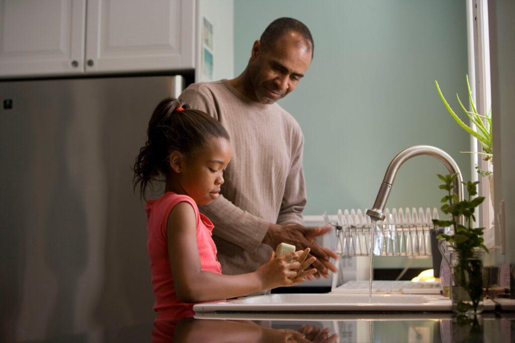 A father and daughter wash their hands in the kitchen sink.