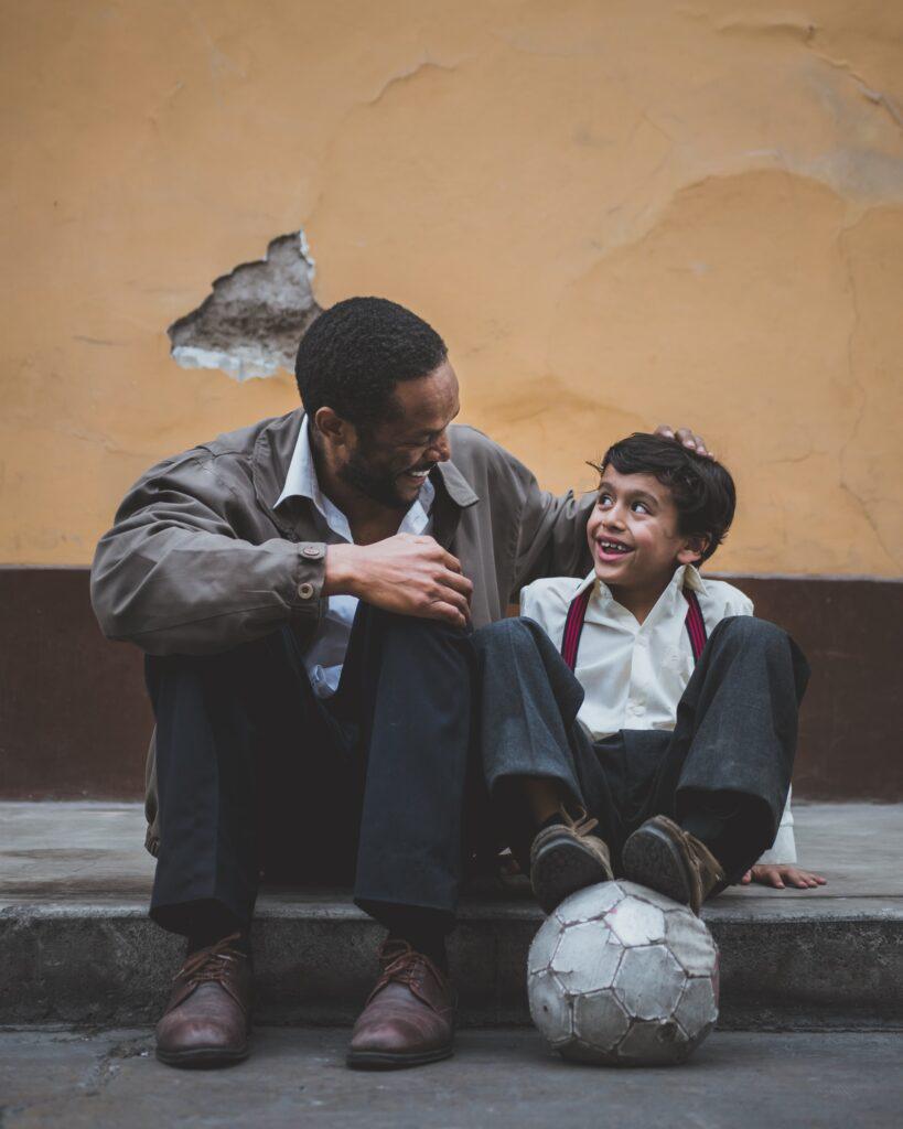A father and son have a casual conversation while sitting on a curb. The boy looks like he just came from school and has a football.