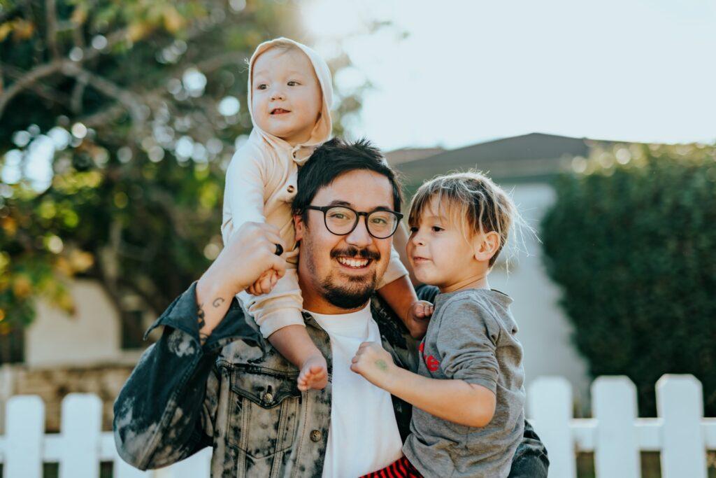 A dad with one child on his shoulder sand the other in his arms.